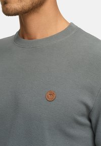 Grå ribbet sweatshirt med rund halsudsk�æring og en struktureret overflade. Har et cirkulært brunt lædermærke på brystet med brandspecifikationer.