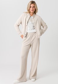 Beige jas met drukknoopsluiting en zijzakken, gecombineerd met bijpassende beige broek. Soepele textuur en een ontspannen pasvorm. Witte sneakers maken de look compleet.