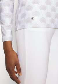 Vit långärmad topp med ett ljusgrått snäckmönster, tillsammans med släta vita leggings. En liten logodetalj på fållen av toppen.