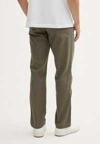 Homme portant un pantalon droit vert olive, des baskets blanches et une chemise blanche, debout devant un fond clair uni.