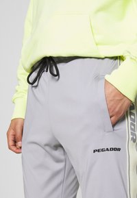 Grå joggerbyxor med elastisk midja, svart dragkedja, sidofickor och en tryckt "PEGADOR"-logotyp på låret. Ytan är slät.