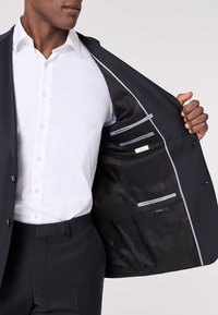 Completo scuro su misura con una camicia bianca. L'interno della giacca presenta una fodera nera testurizzata, tasche con dettagli grigio chiaro e etichette.