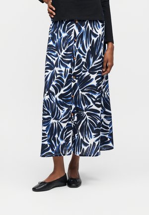 Žena v modro-čiernej midi sukni s listovým vzorom a prednými gombíkmi, čiernom topu a čiernych balerínach, stojaca s jednou rukou na boku.