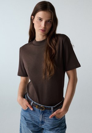 Junge Frau mit langen braunen Haaren, die ein dunkelbraunes kurzärmeliges Oberteil und blaue Jeans mit braunem Gürtel trägt, steht mit den Händen in den Taschen.