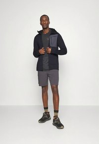 Svart huva jacka med en grå bröstficka, kombinerad med en mörkgrå t-shirt och kolgråa shorts. Svarta sportskor och strumpor fulländar outfiten.