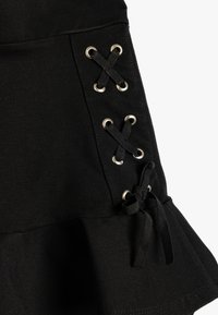 Jupe noire avec un design à lacets comportant des œillets argentés et des lacets noirs. L'ourlet est évasé avec un bord froncé. Tissu doux et texturé.