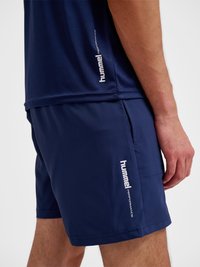 Mężczyzna w granatowych sportowych szortach hummel Performance i pasującej koszulce, stojący z ręką spoczywającą wzdłuż ciała.