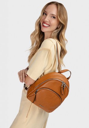 Mujer sonriente con cabello largo y ondulado rubio, vistiendo un conjunto beige claro y llevando una mochila de cuero marrón medio en el hombro.