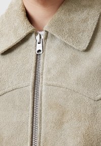 Suedesignerjacka i ljusbeige med spetsig krage och central silverdragkedja. Texturerad yta med subtila sömnadsdetaljer.