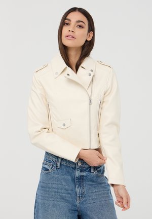 Giacca biker cropped in eco-pelle color crema con colletto a revers, chiusura zip e dettagli con bottoni a pressione. Abbinata a jeans blu a vita alta.