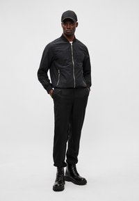 Schwarze Bomberjacke mit Reißverschluss, zwei Seitentaschen und gerippten Bündchen, kombiniert mit schwarzen Hosen und klobigen schwarzen Schnürstiefeln.