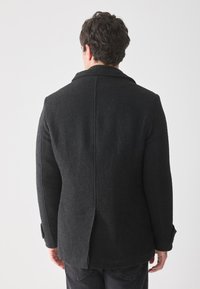 Hombre visto de espaldas vistiendo un abrigo de lana texturizada negro y pantalones oscuros, contra un fondo claro y liso.