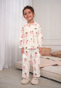 Ensemble de pyjama blanc avec motifs de ballerines roses. Haut à manches longues avec col et fermeture à boutons. Pantalon ample. Chaussons doux et confortables.