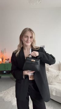 Jeune femme en blazer et pantalon noirs tenant un petit sac à main noir, debout dans un salon confortable à la décoration neutre.