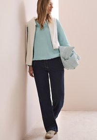 Maglione lungo a maniche lunghe blu chiaro con texture, maglione corto bianco appoggiato sulle spalle, jeans blu scuro, in mano una giacca trapuntata blu chiaro, sneakers beige.