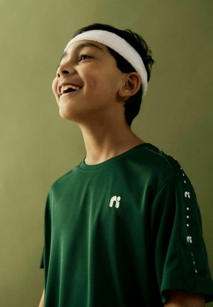 Niño sonriendo con una cinta deportiva blanca en la cabeza y una camiseta deportiva verde oscuro con un logo blanco en el pecho y la manga, contra un fondo verde.