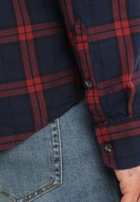 Flanellen overhemd met een marineblauw en rood geruit patroon, knoopsluiting bij de pols en een afgeronde zoom, gecombineerd met licht denim jeans.