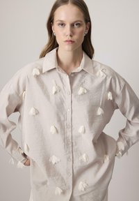 Camisa de color beige claro con un corte relajado, que presenta aplicaciones florales blancas, un frente abotonado y un cuello clásico. Tejido texturizado.