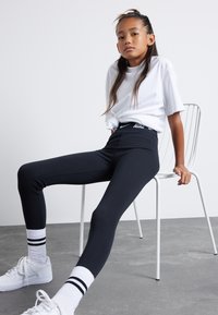 Black high-waisted leggings, smooth texture, with white Nike logo. Paired with a white t-shirt and white sneakers with black stripes on socks.