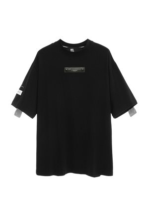 Camiseta negra de gran tamaño con manga corta y cuello redondo, pequeño parche rectangular con texto en el pecho y etiquetas blancas en las mangas.