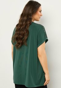 Top verde a maniche corte in un fit morbido con una texture liscia, caratterizzato da dettagli in paillettes sulle spalle e orlo arrotondato.