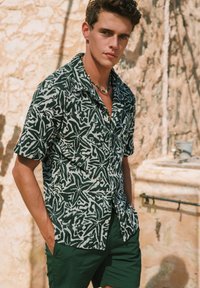 Camicia a maniche corte di colore verde scuro con un motivo di foglie astratte bianche; realizzata in tessuto leggero. Abbinata a pantaloni corto verde coordinati.