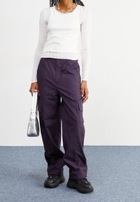 Bílý top s dlouhým rukávem v kombinaci s tmavě fialovými cargo kalhotami se bočními kapsami a černými robustními teniskami. Stříbrná kabelka dodává kontrast.