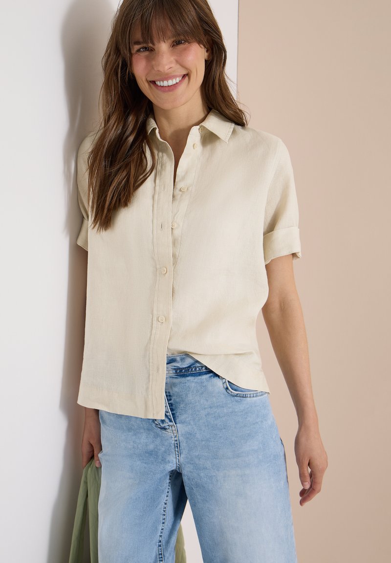 Femme souriante aux longs cheveux bruns, portant une chemise beige à manches courtes boutonnée et un jean bleu clair, tenant un tissu vert dans une main.