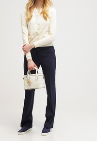 Cardigan crema, pantalones azul marino y zapatos azul marino. Sosteniendo un pequeño bolso blanco con herrajes dorados y una etiqueta. Diseño simple y limpio.