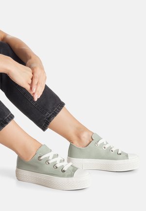 Persona que lleva zapatillas de lona de corte bajo de color verde claro con suelas blancas gruesas y cordones, sentada con las manos apoyadas sobre pantalones negros.