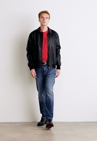 Črna usnjena bomberska jakna nad rdečo majico, modri kavbojki in črnimi čevlji. Enostavna podlaga s figurico, ki stoji v nevtralnem položaju.