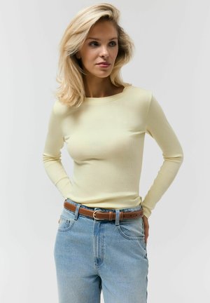 Blondynka o średniej długości włosach, ubrana w jasnżółty top z długim rękawem, wpuszczony w spodnie wysokiego stanu w kolorze niebieskim, z brązowym paskiem.