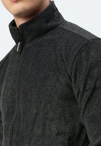 Schwarze Fleecejacke mit hohem Kragen, durchgehendem Reißverschluss und strukturiertem Material. Hat ein Logo am Zipper und eignet sich für einen regulären Schnitt.