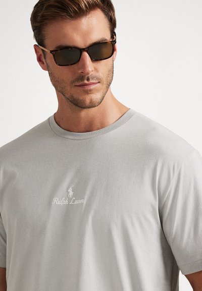 Mann som bruker solbriller og en lys grå Ralph Lauren t-skjorte med en liten logo på brystet, vendt litt mot venstre.