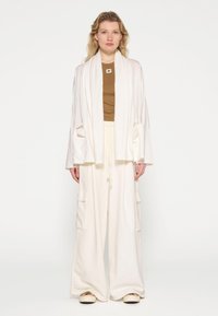Witte oversized sjaal en losse witte broek met zakken, gelaagd over een bruin topje. Glad materiaal en relaxte pasvorm, geschikt voor casual gebruik.