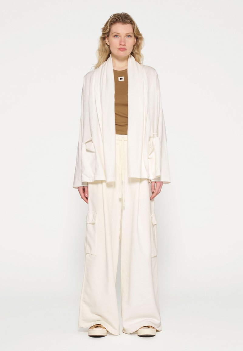 Witte oversized sjaal en losse witte broek met zakken, gelaagd over een bruin topje. Glad materiaal en relaxte pasvorm, geschikt voor casual gebruik.