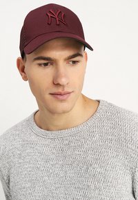 Cappello bordeaux con logo ricamato, fronte strutturato e lati in rete. Abbinato a un maglione grigio chiaro di tessuto testurizzato.
