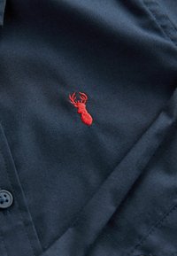 Tengerészkék ing, bal oldalon piros hímzett szarvas emblémával, texturált anyaggal és gombos részletekkel.