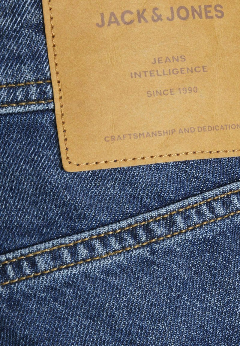 Denim w głębokim niebieskim kolorze, z brązową skórzaną metką z wytłoczonym tekstem. Gładka tekstura z widocznymi detalami przeszycia.