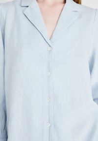 Camicia di lino azzurra chiara con colletto a scollo a V, bottoni frontali e tessuto strutturato. Il design semplice enfatizza la morbidezza e la traspirabilità.