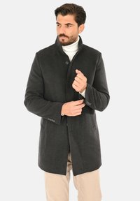 Cappotto in lana grigio scuro con collo alto, chiusura con bottoni e tasche laterali. La texture liscia e la vestibilità sagomata esaltano la sua forma strutturata.