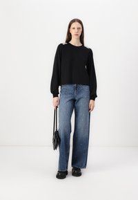 Μαύρη μπλούζα με μακριά μανίκια και διακοσμητικές ασημένιες λεπτομέρειες στους ώμους, συνδυασμένη με παντελόνι τζιν σε φαρδιά γραμμή και λευκές μπίλιες ως διακόσμηση.