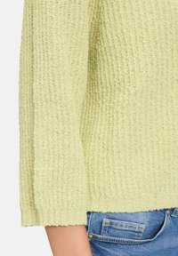 Hellgrüner, gestrickter Pullover mit gerippter Textur und drei Viertellängenärmeln, kombiniert mit blauen Jeans, die das Detail der Manschette zeigen.
