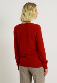 Pull en tricot rouge avec des manches longues et un col rond, associé à un pantalon à motifs beige et noir. Texture douce, coupe décontractée.