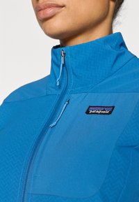 Μπλε μπουφάν με ψηλό γιακά, κλείσιμο με φερμουάρ και υφή με σχέδιο ψαροκόκκαλο. Διαθέτει λογότυπο της Patagonia στην πάνω αριστερή πλευρά.