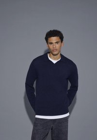 Marineblauer Pullover mit V-Ausschnitt und kurzem Kragen, gefertigt aus weichem Stoff. Über einem weißen Hemd getragen, kombiniert mit dunkelgrauen Hosen.