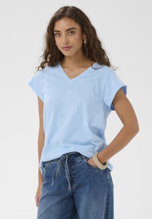 Žena s kučeravými hnedými vlasmi má na sebe svetlomodrý top s výstrihom do V a krátkymi rukávmi a modré rifle so sťahovacou šnúrkou, pózuje s rukou vrecku.