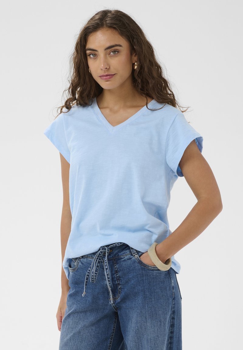 Sieviete ar cirtainiem brūniem matiem nēsā gaiši zilu V-veida izgriezuma īsām piedurknēm topu un zilas džinsas ar auklu, pozējot ar roku kabatā.