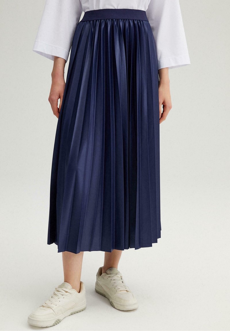 Gonna a pieghe blu navy con una vita aderente, realizzata in un tessuto liscio e lucido. Abbinata a sneakers bianche per un look casual.