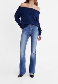 Granatowy sweter bez ramiączek z prążkowanym dekoltem, w połączeniu z jasnoniebieskimi jeansami w dzwony z bocznymi rozcięciami i ciemnymi obcasami.